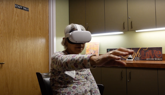 An older woman wearing a white virtual reality headset sits in a chair, extending her arm forward and interacting with a virtual environment.